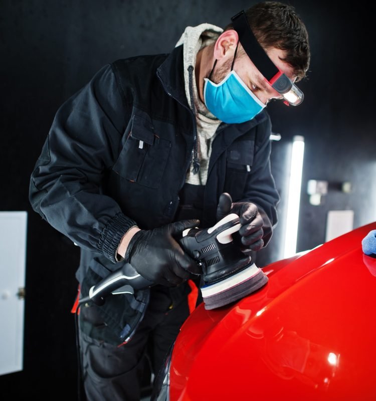 Car detailing concept. Man in face mask with orbital polisher in repair shop polishing orange suv car.